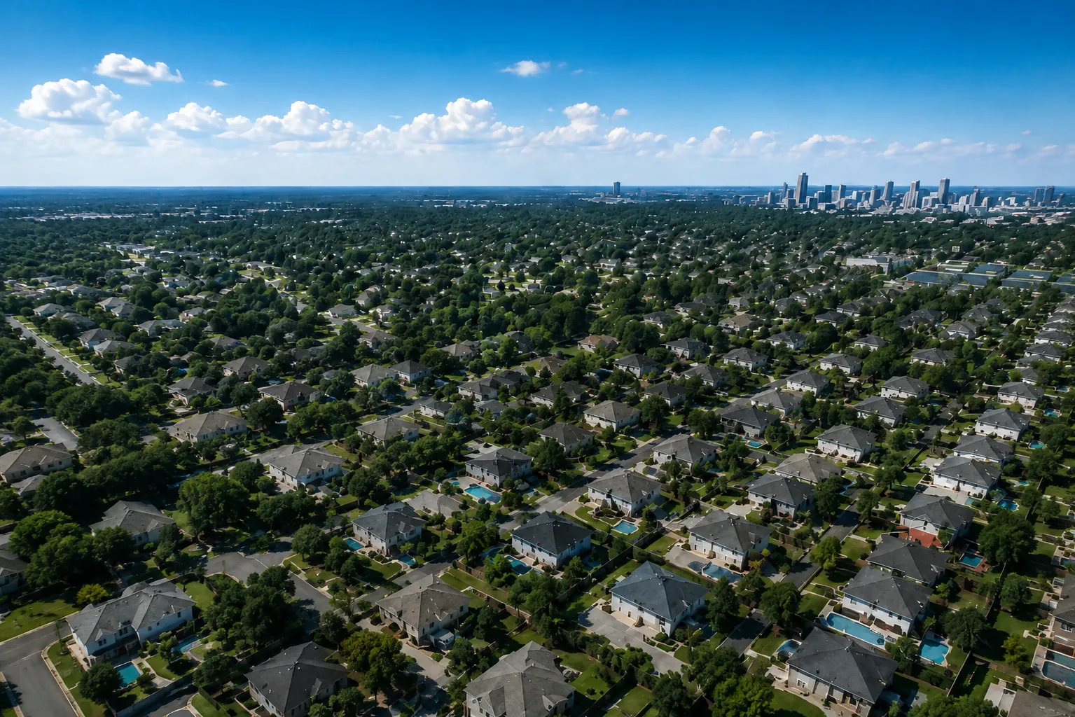 Aerial view of a tree-lined residential neighborhood with lawns, driveways, and pools, a city skyline on the horizon